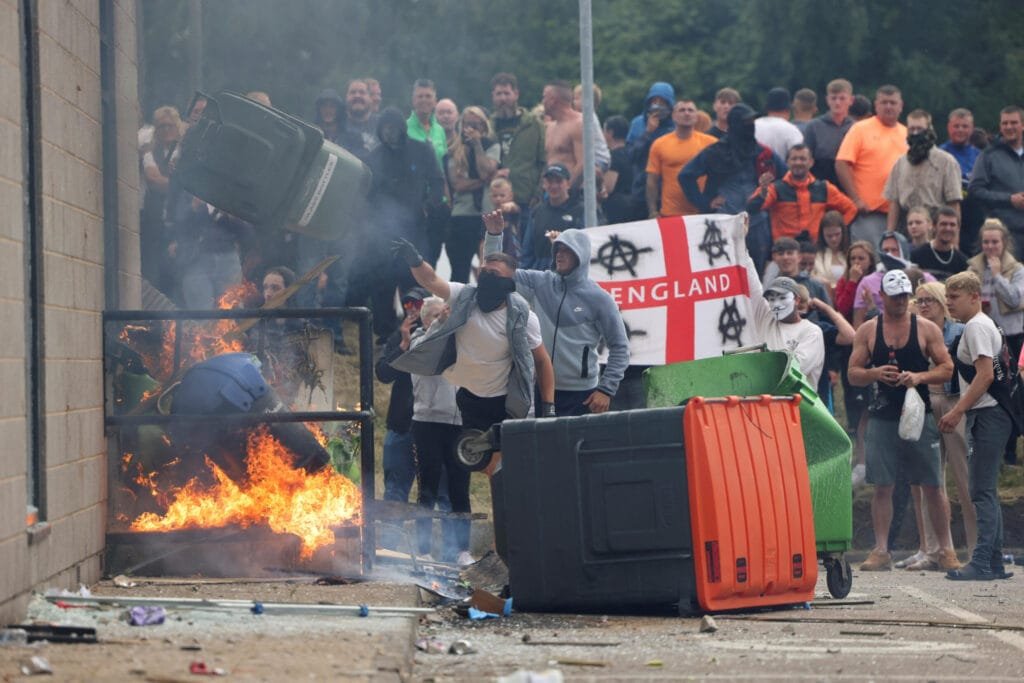 Disturbios en el Reino Unido protagonizados por manifestantes racistas anti-inmigración. La imagen muestra a un grupo de personas enmascaradas y encapuchadas, arrojando contenedores de basura a un fuego mientras otros observan con una bandera de Inglaterra marcada con símbolos anarquistas. La violencia se desató en medio de protestas contra la inmigración, reflejando tensiones sociales crecientes en el país.
