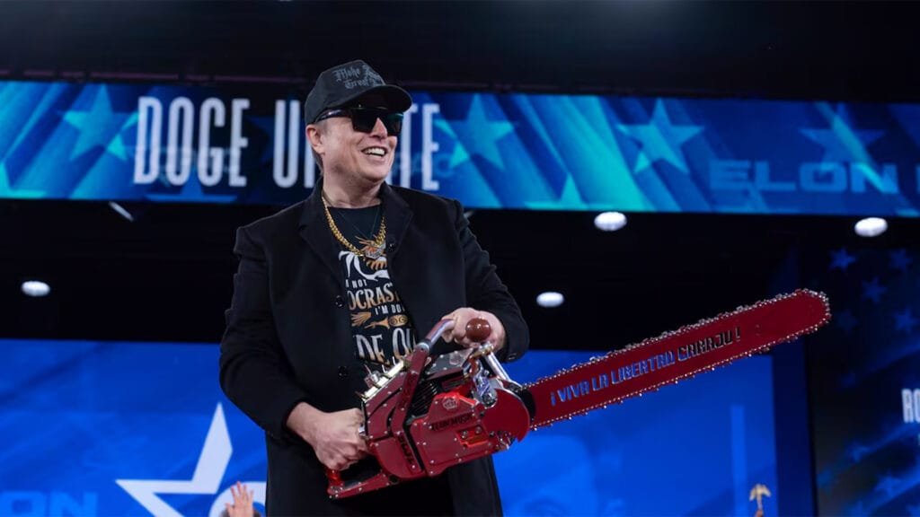 Elon Musk en un escenario. Tiene puesta una gorra y unos lentes de sol y sonríe mientras sostiene una motosierra roja en la que puede leerse "Viva la libertad carajo".